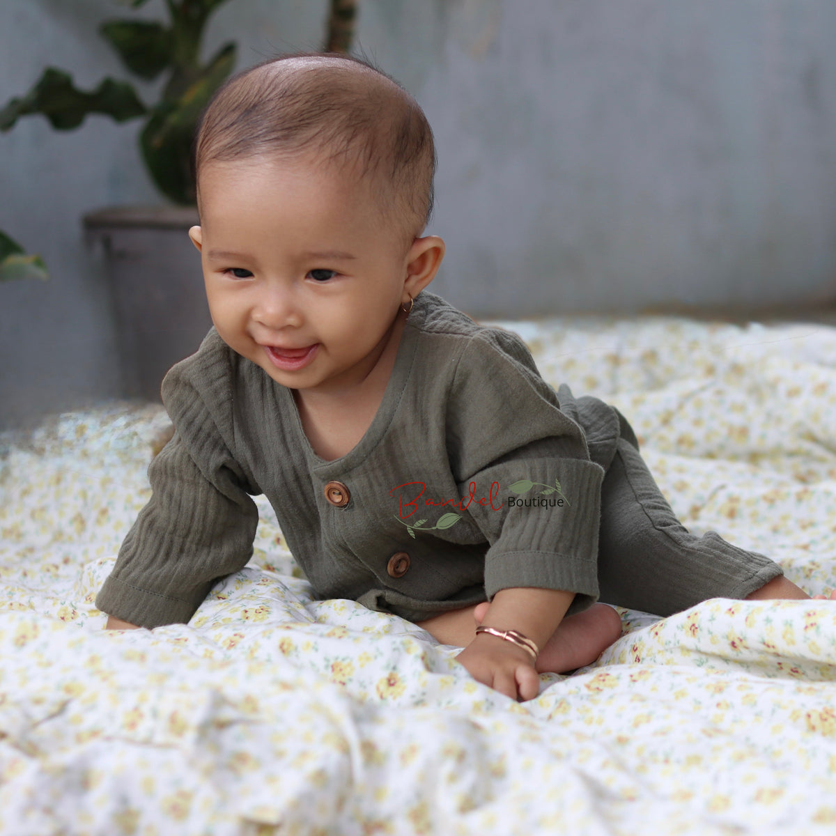 A baby wearing a ribbed long-sleeve romper in a muted olive green, crawling on a white and yellow floral patterned blanket in a softly lit indoor setting.
