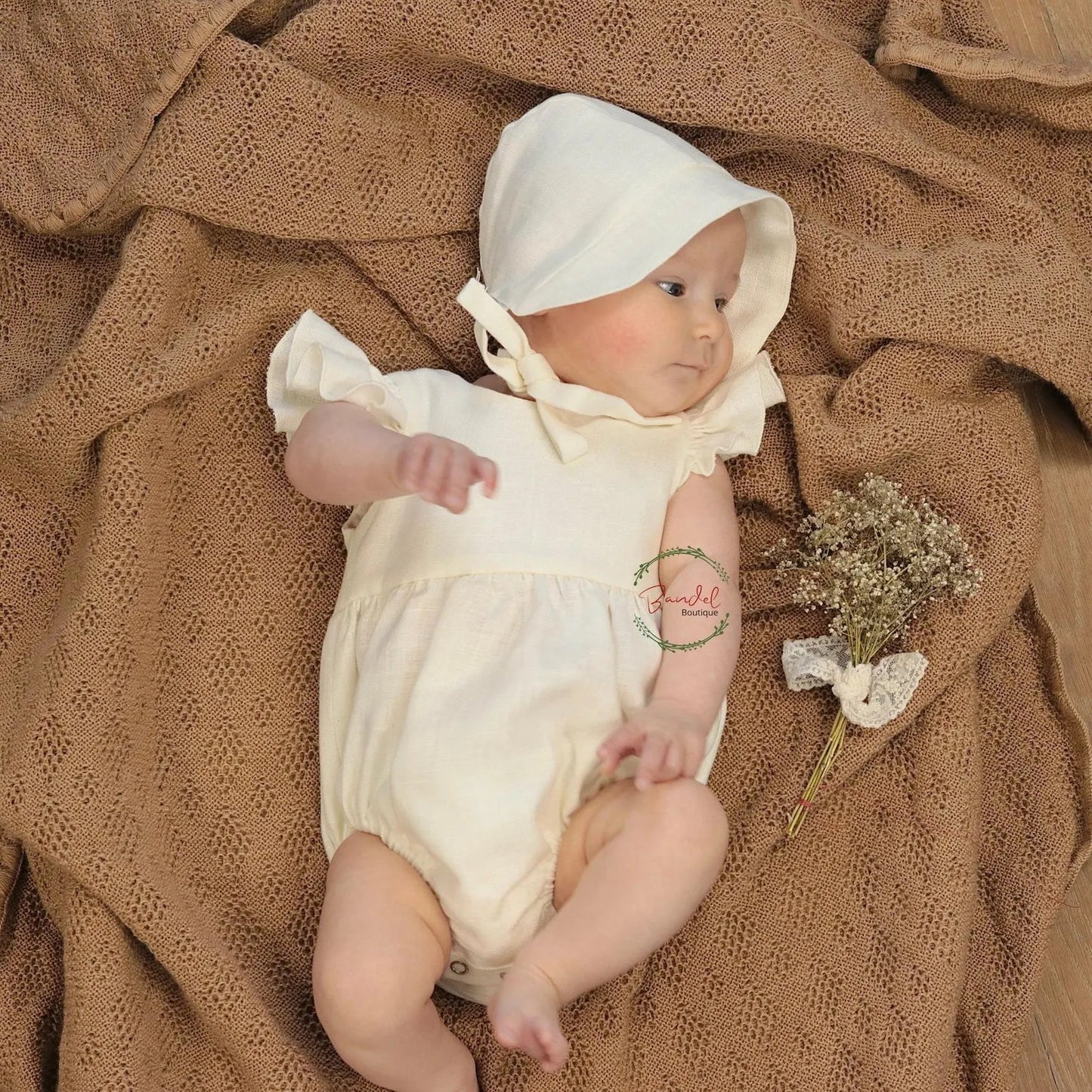 Baby in a white outfit and bonnet lying on a brown textured blanket with a small bouquet of flowers.
