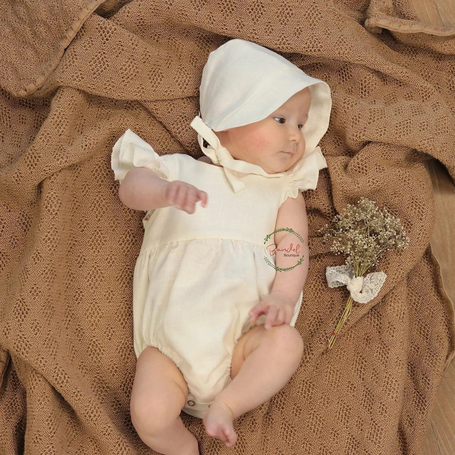 Baby in a white outfit and bonnet lying on a brown textured blanket with a small bouquet of flowers.