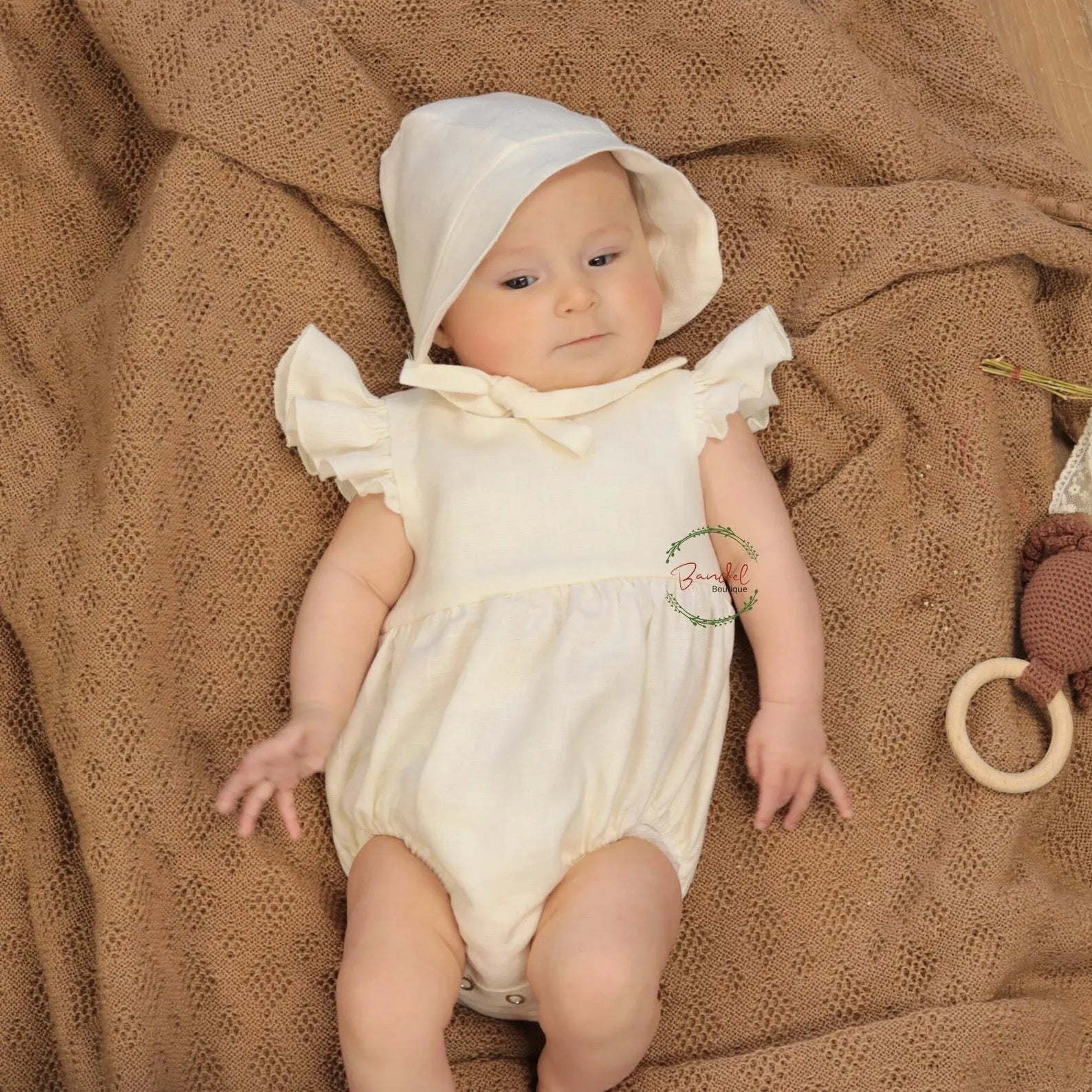 Baby in a white outfit and bonnet lying on a brown textured blanket.
