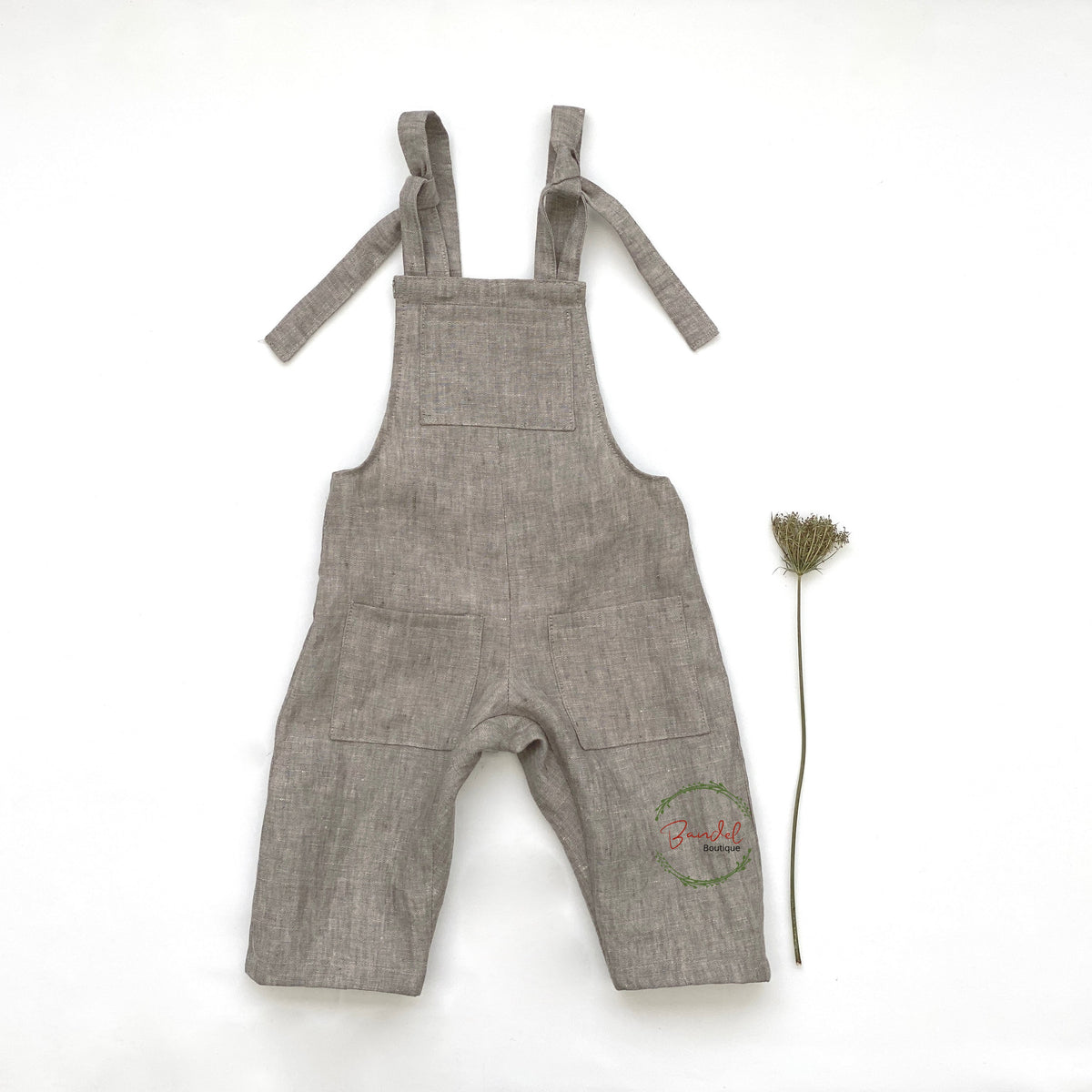A pair of khaki linen dungarees for children, with adjustable straps and three pockets, displayed with a small green plant.
