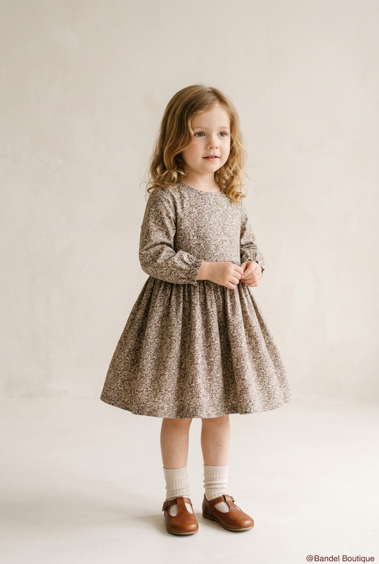 A young girl with wavy light brown hair stands in a studio wearing a long-sleeve, knee-length dress with a dense small-scale floral or abstract print in warm taupe, beige, and brown tones. She is styled with cream socks and brown leather Mary Jane shoes against a warm, textured off-white background.