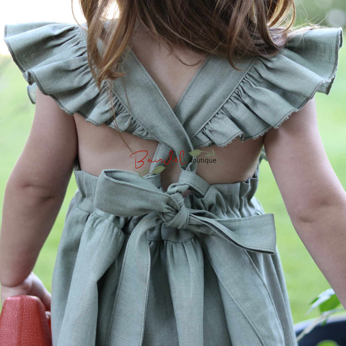 A child wearing a sage green ruffled dress, shown from behind in an outdoor garden setting.