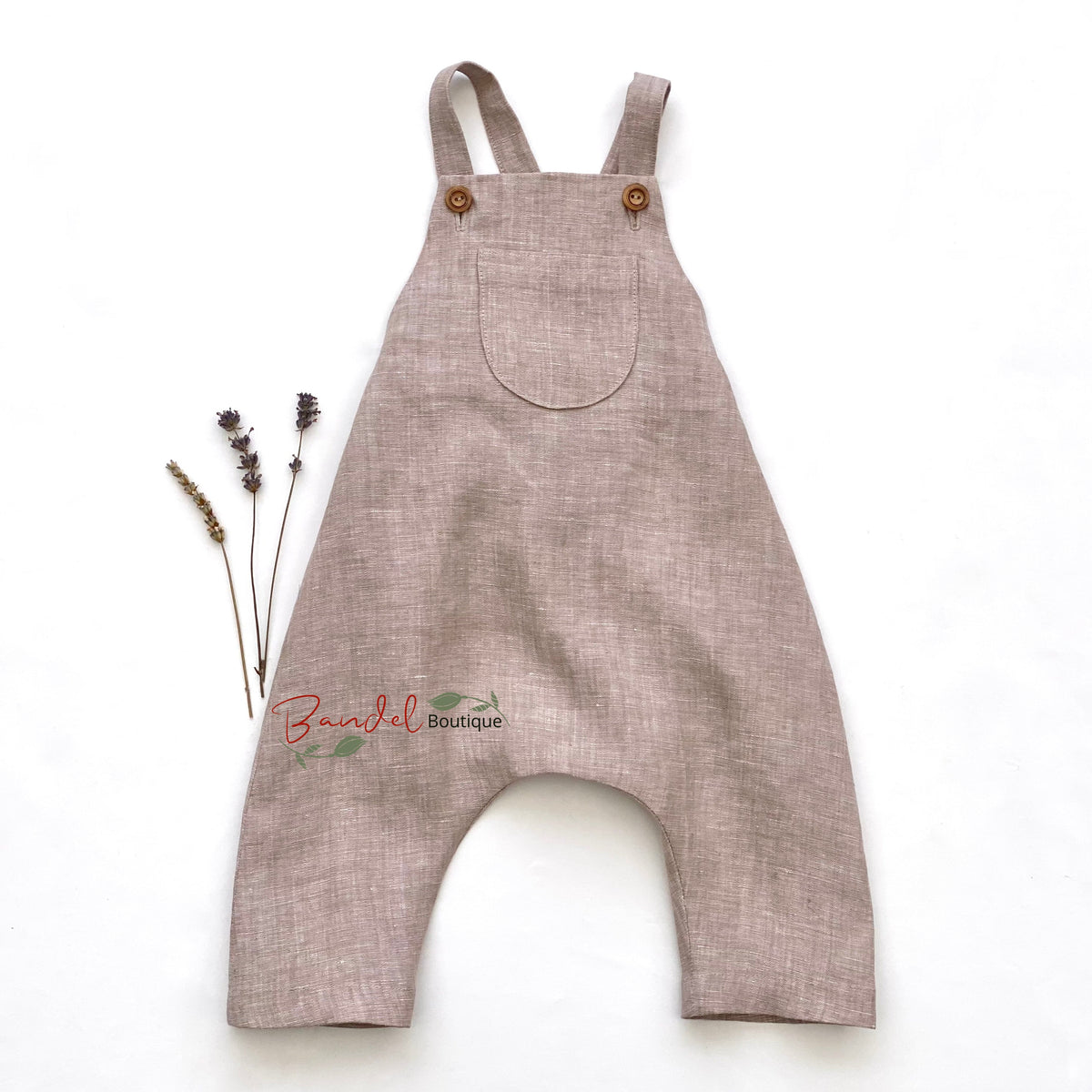 A light brown linen romper with wide legs and shoulder straps, laid flat on a white background next to dried lavender sprigs.