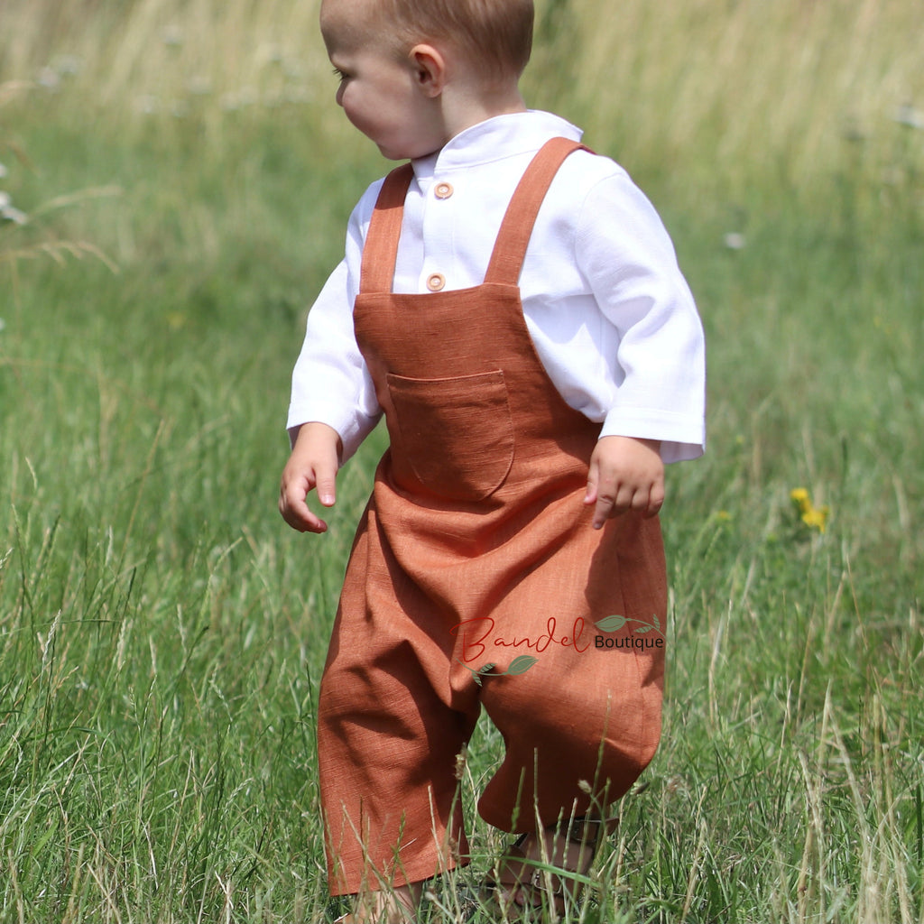 Rust Linen baby romper dungarees feature 3/4 length shorts with a traditional curved front pocket. The elastic back waistband and adjustable tie straps provide a secure fit, and the wooden button closure at the back creates a stylish look.