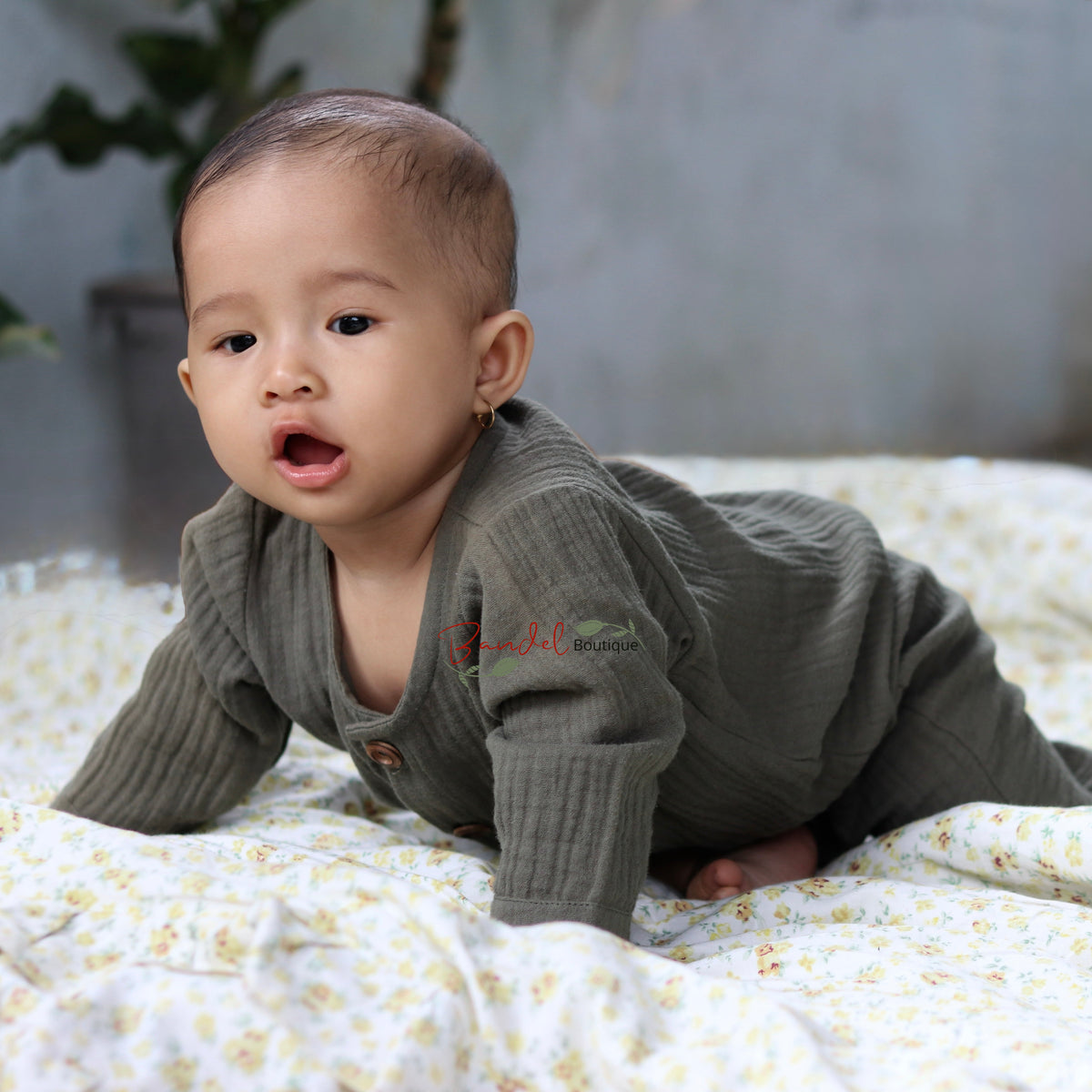 A baby crawling on a white floral blanket, wearing a ribbed long-sleeve romper in a muted olive green color.