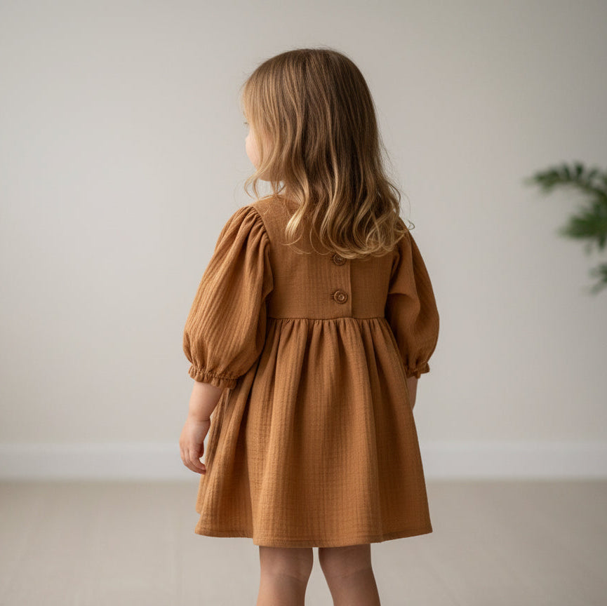 Young girl wearing a brown dress standing against a plain wall.