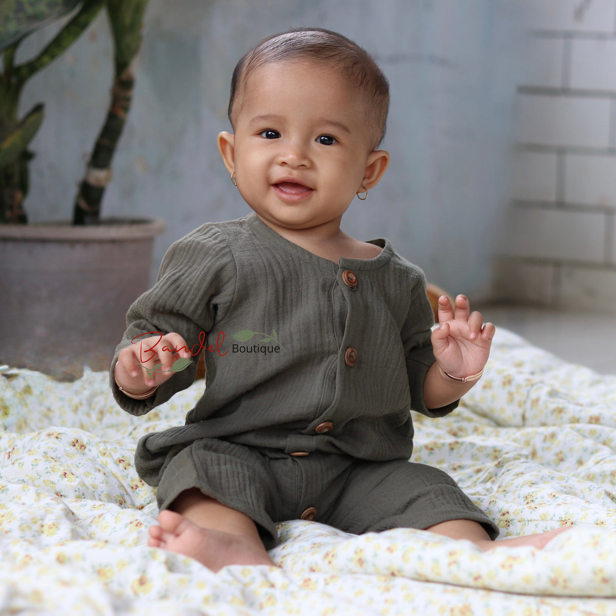 A baby sitting on a white floral blanket in a room, wearing an olive green ribbed long-sleeve romper with brown buttons.