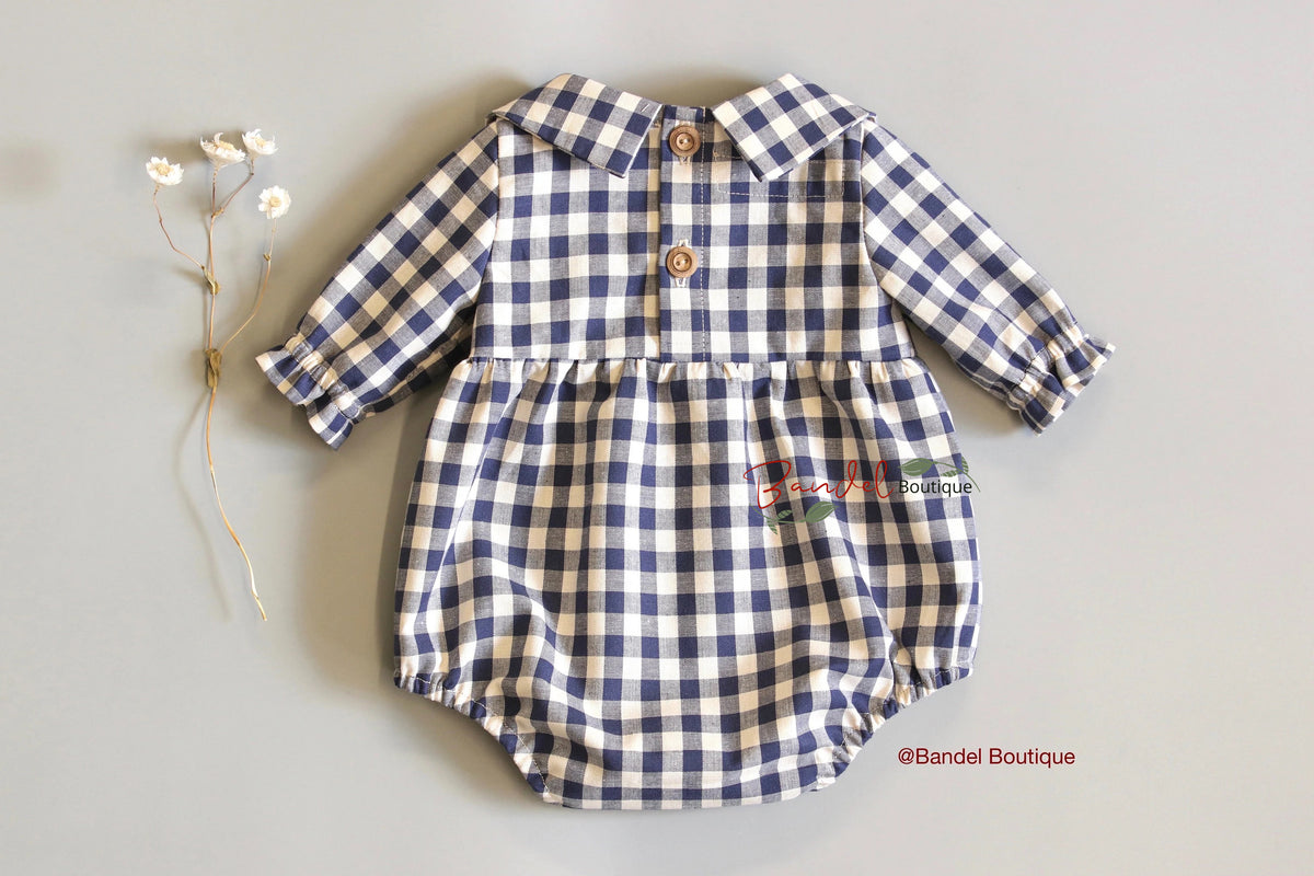 A baby romper with a navy blue and cream gingham check pattern, featuring a pointed collar, three wooden buttons, and a ruffled waist seam. It is displayed flat on a light gray background next to a small sprig of dried white flowers.