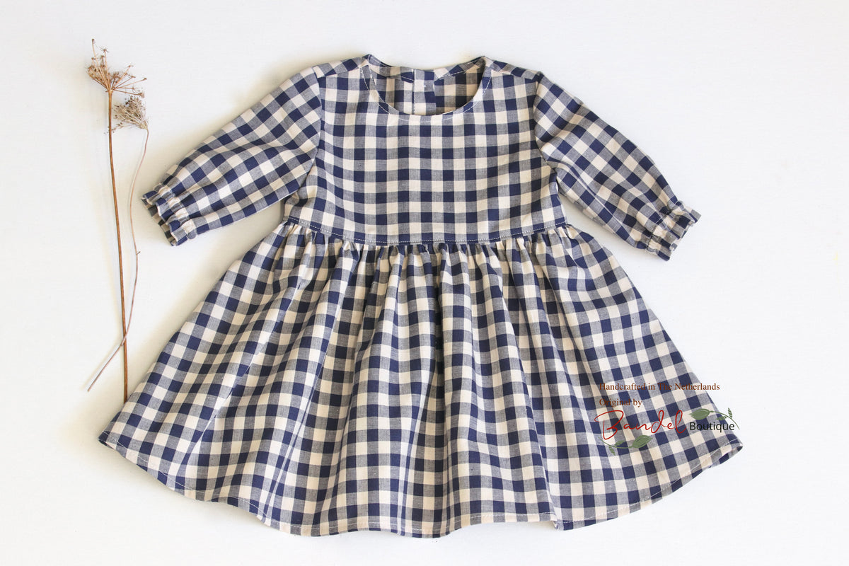 A toddler girl's long-sleeve dress with a navy blue and cream gingham check pattern, laid flat on a white background next to a dried flower stem.