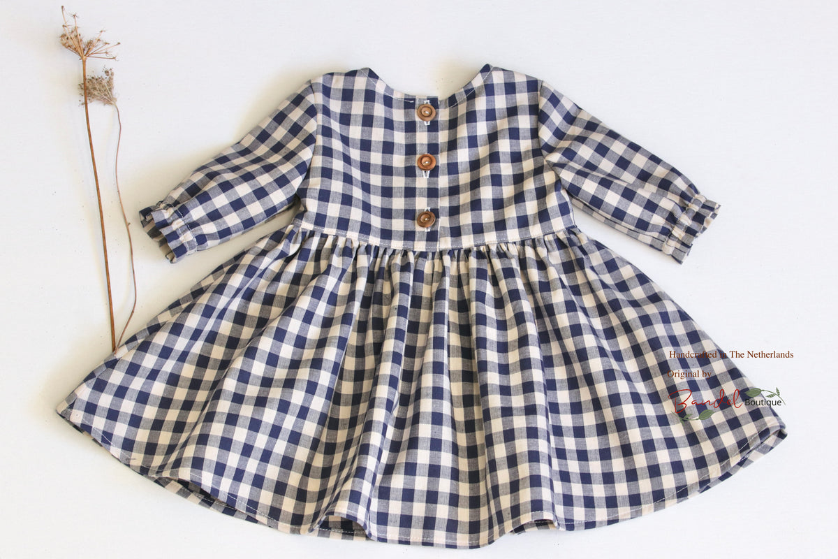 A long-sleeve baby dress with a navy and cream gingham check pattern, featuring three brown buttons on the chest and a gathered waist, laid flat on a white background next to a dried flower stem.