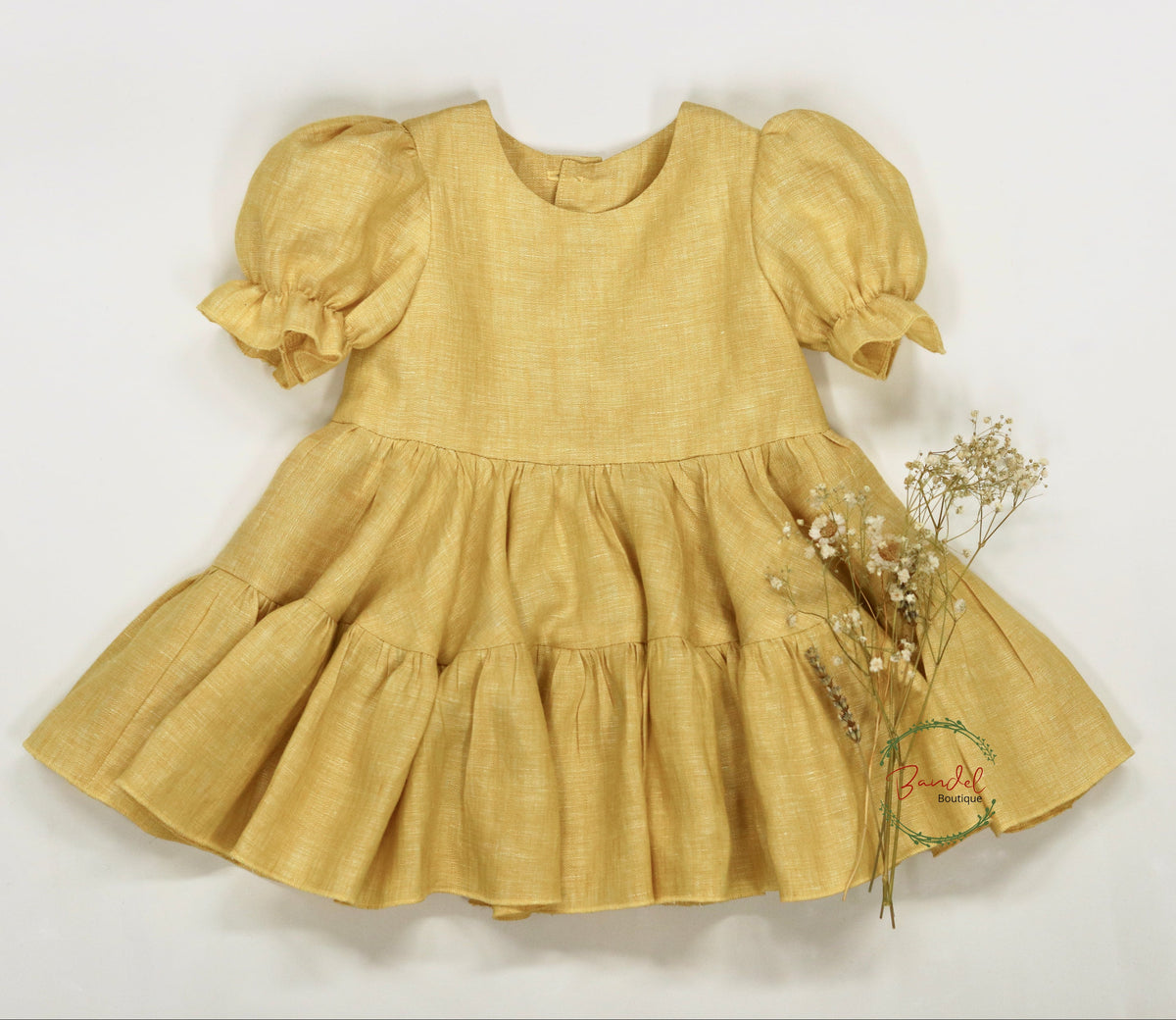 A yellow linen baby dress with short puff sleeves and a tiered ruffled skirt, laid flat on a white background next to dried baby's breath flowers.