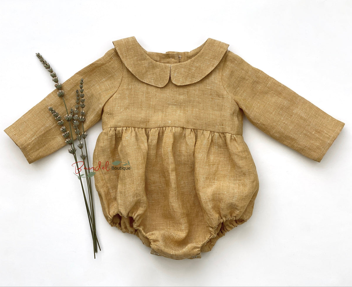 An ocher yellow linen romper for babies, with long sleeves, a Peter Pan collar, wooden buttons at the back, and elastic at the leg. It is displayed with a small bunch of dried plants, and is laid out flat on a white background.