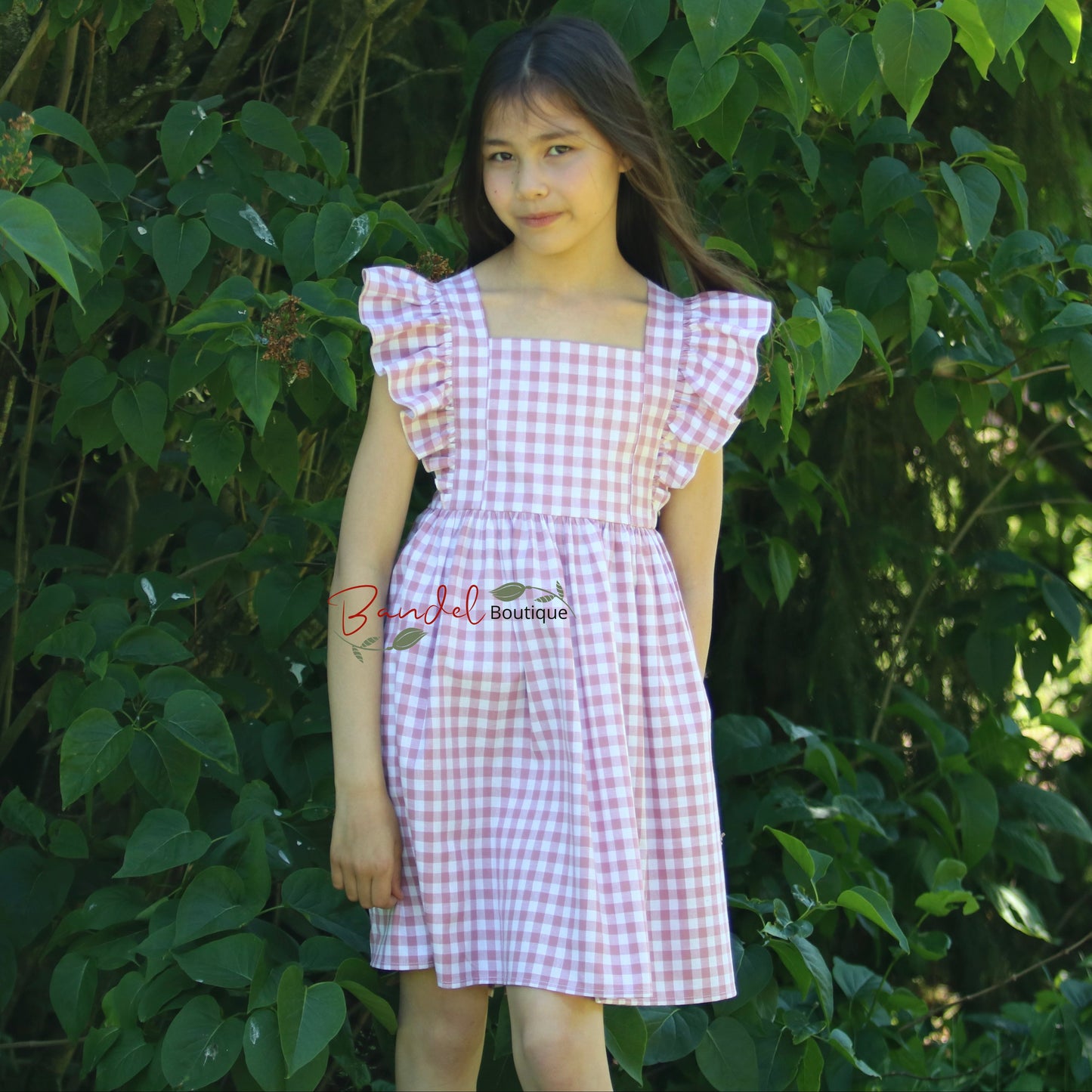 Young girl in a checkered dress standing in front of green foliage