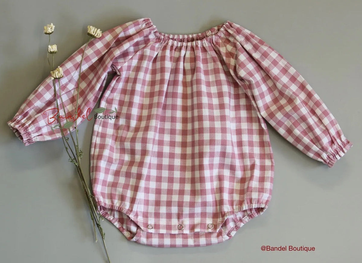 A pink and white gingham long-sleeve baby romper with an elasticized neckline and snap closures at the crotch, laid flat on a neutral gray background next to dried white flowers. A watermark for 'Bandel Boutique' is visible.