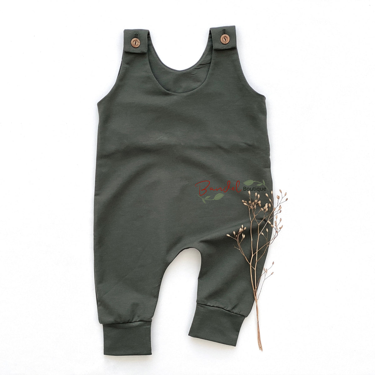 An olive green baby romper laid flat on a white background, styled with a small sprig of dried grass. The romper features adjustable shoulder straps with wooden buttons and a small red embroidered logo on the chest.