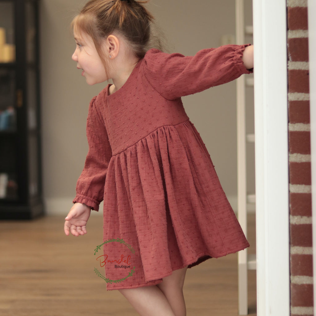 Young girl in a red dress standing in a room with wooden floor and brick wall.