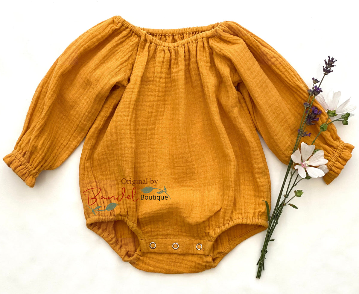 A mustard yellow crinkled gauze baby bodysuit with long puff sleeves and snap closures at the crotch, laid flat on a white background next to small white and purple flowers. A watermark reading 'Original by Bella Boutique' is visible on the front.