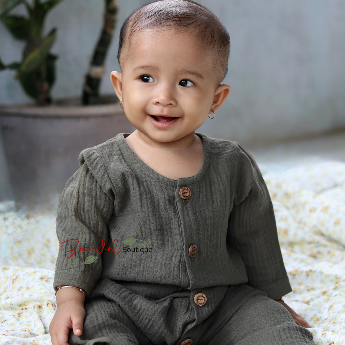 A smiling baby wearing a ribbed olive green long-sleeve romper with brown buttons, sitting on a white floral blanket in a softly lit indoor setting.