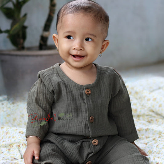 A smiling baby wearing a ribbed olive green long-sleeve romper with brown buttons, sitting on a white floral blanket in a softly lit indoor setting.