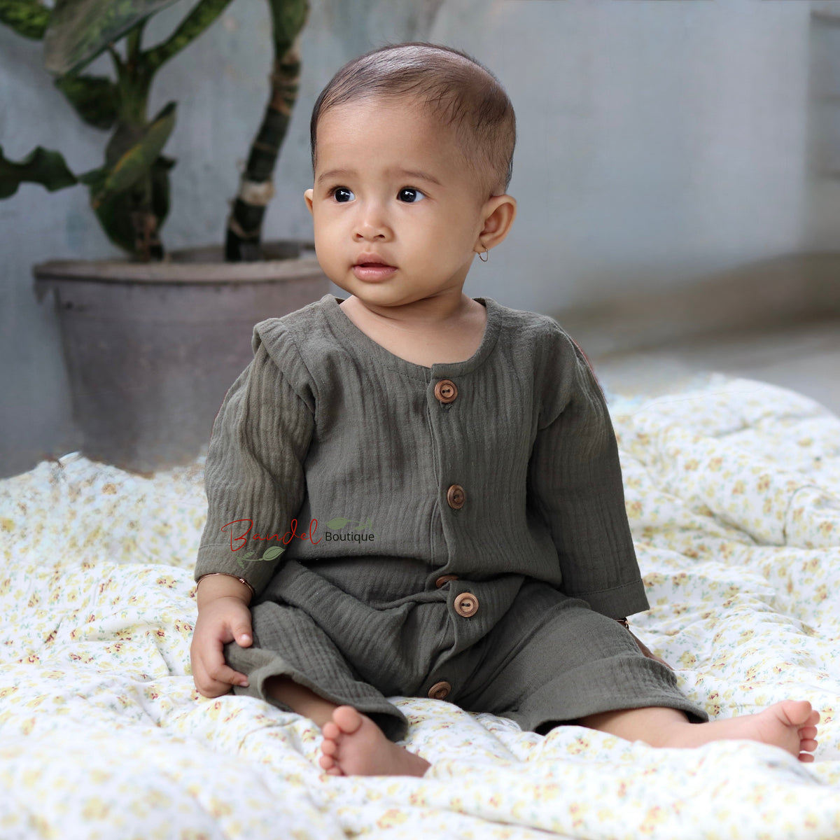 A baby sitting on a white and yellow floral blanket, wearing a ribbed olive green long-sleeve romper with brown buttons.