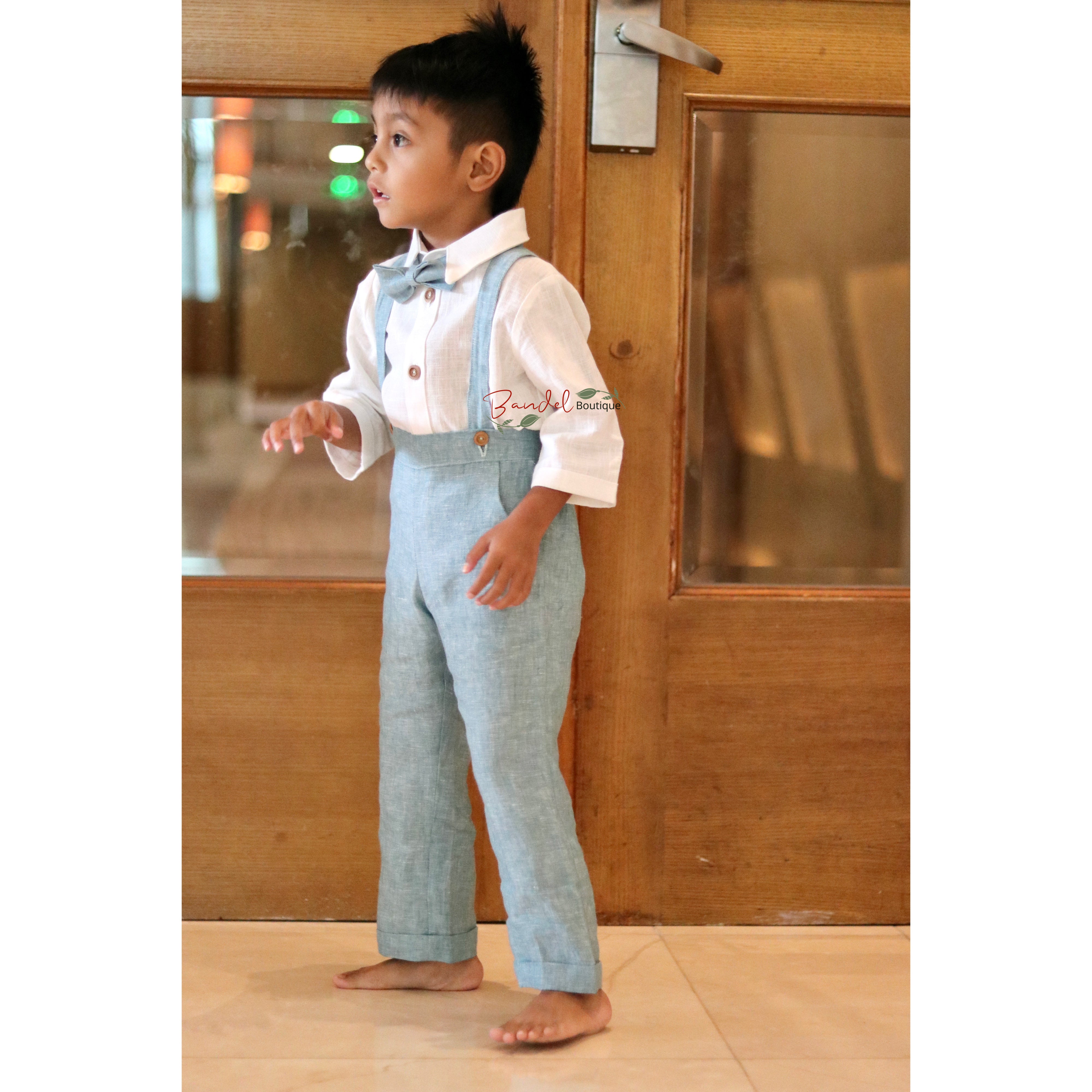 Child wearing a white shirt with a bow tie and light blue pants standing in front of a wooden door.
