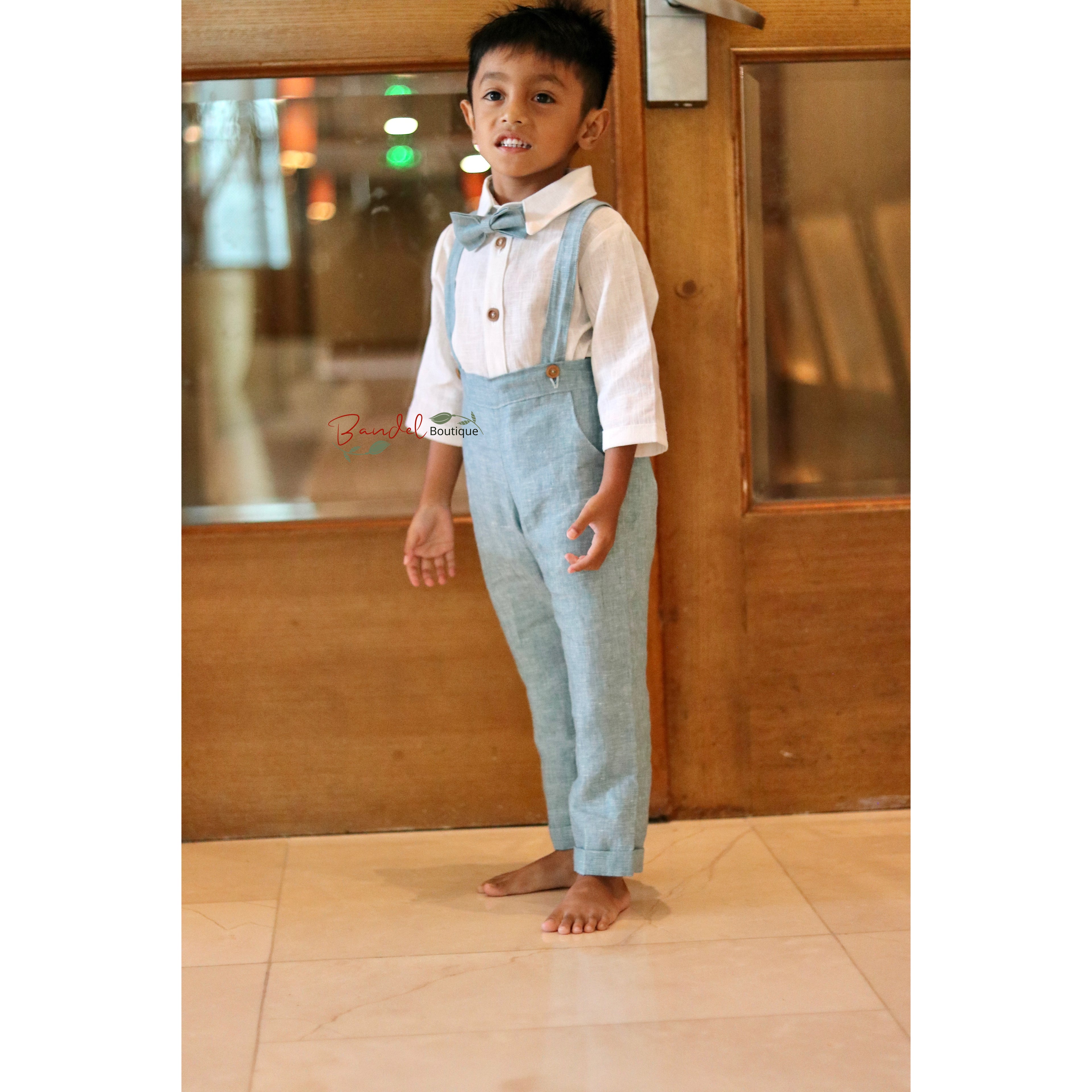 Child wearing a white shirt, blue suspenders, and a bow tie standing in front of a wooden wall.