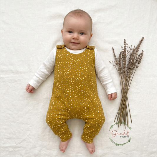 A baby lying on a white fabric background wearing a mustard yellow and white polka dot overall romper over a white long-sleeve shirt, with dried lavender stems placed to the right and a 'Brindel Boutique' watermark near the bottom right.