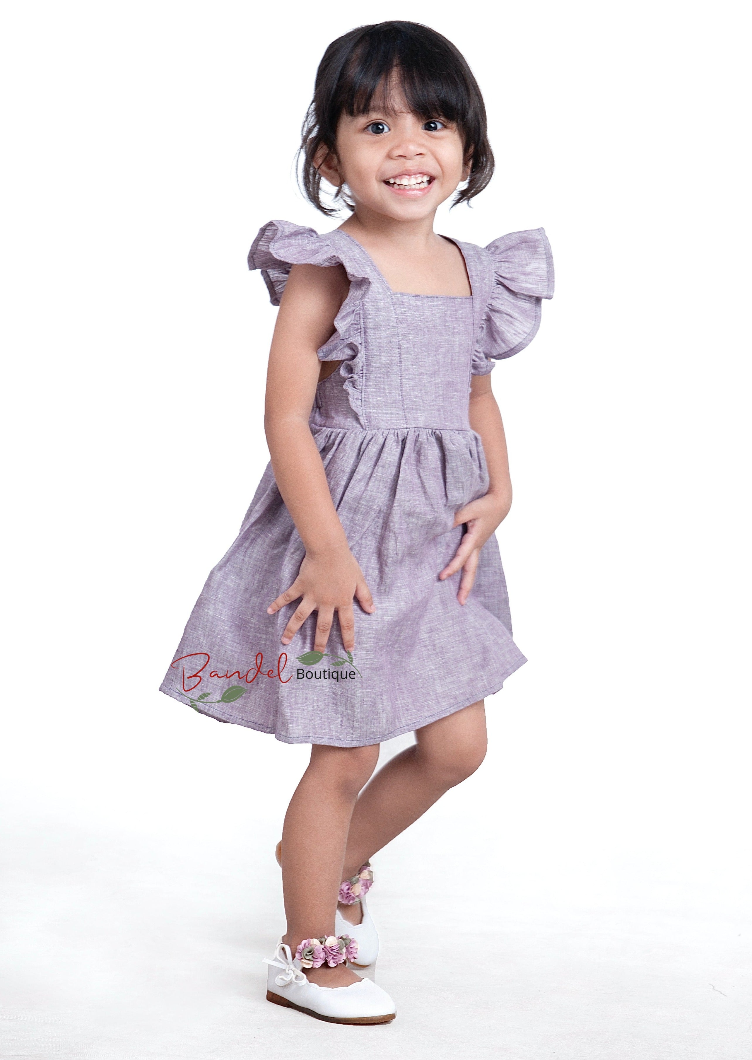 A young girl wearing a purple linen flower girl dress with ruffled sleeves and a gathered skirt.