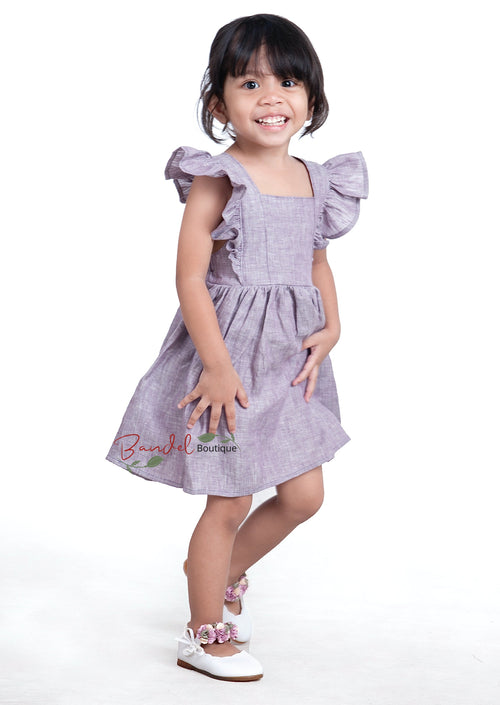 A young girl wearing a purple linen flower girl dress with ruffled sleeves and a gathered skirt.
