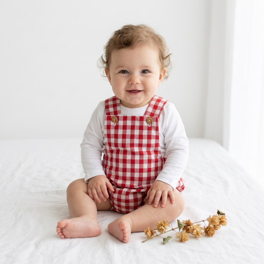 A baby sitting on a white bed wearing a red and white gingham check overalls romper over a white long-sleeve shirt, with a small dried flower sprig in the foreground.