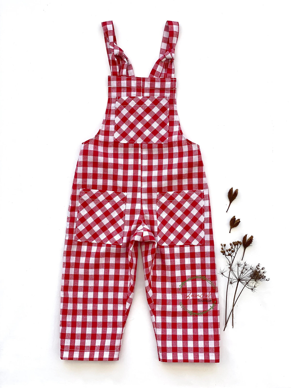 Red and white gingham checkered children's overall jumpsuit with shoulder straps and front pockets, laid flat on a white background next to dried flowers.