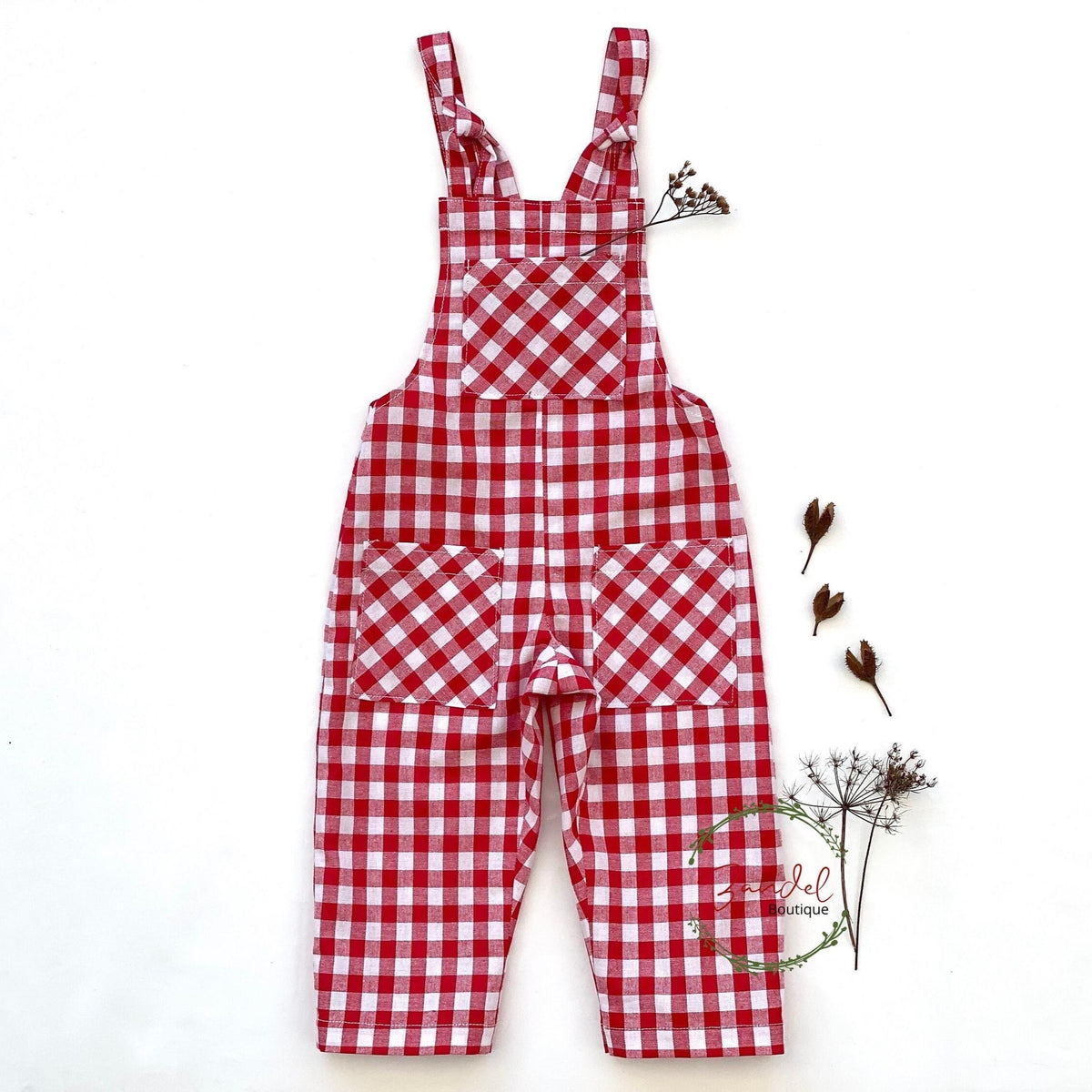 Red and white gingham check bib overalls with front patch pockets, laid flat on a white background with small dried floral props.