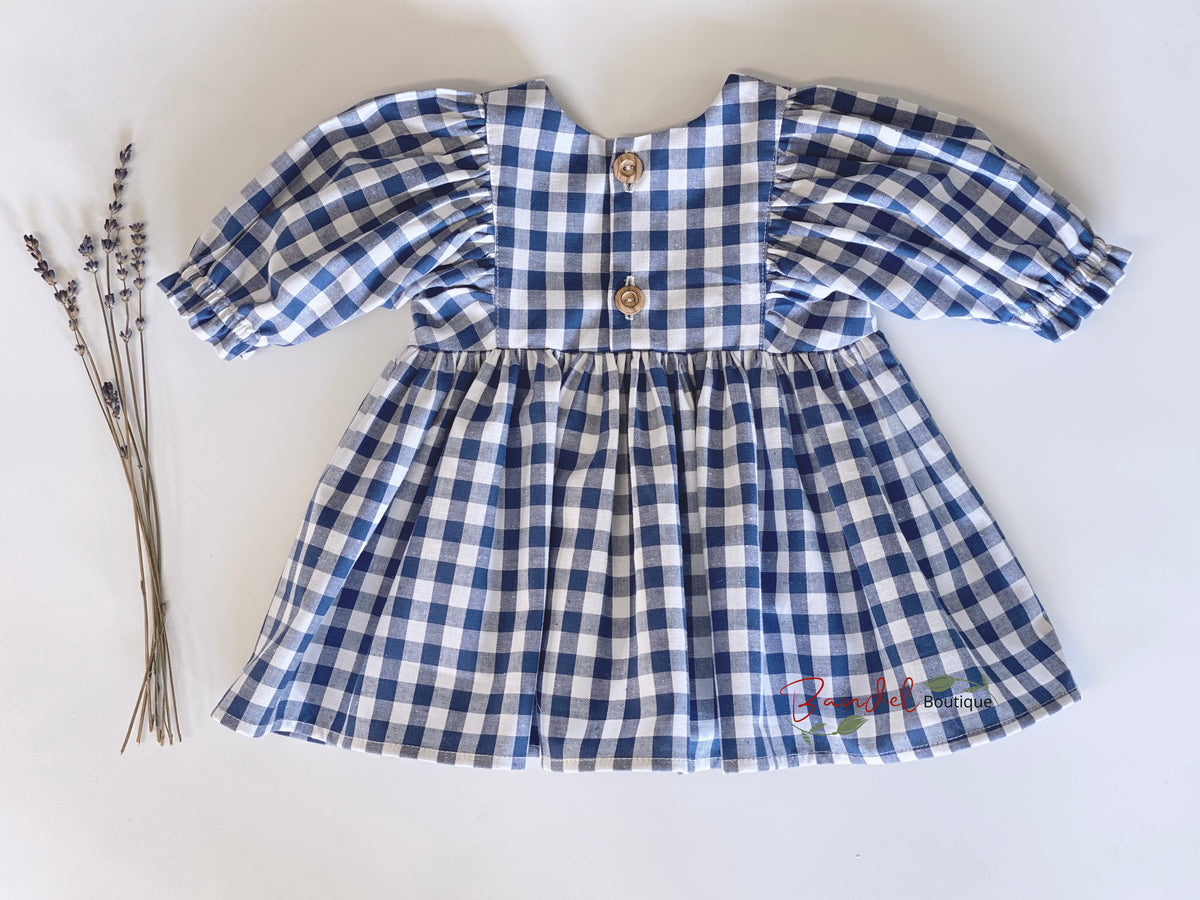 A blue and white gingham dress for toddlers, featuring puff sleeves, a gathered waist, and two wooden buttons on the back, laid flat on a white background next to dried lavender sprigs.