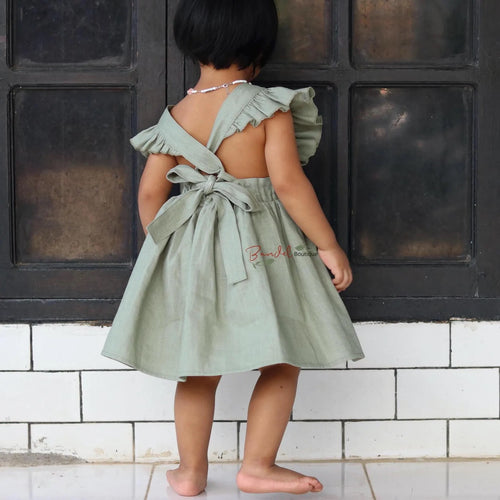 Child wearing a green dress with ruffled sleeves and a bow tie, standing against a black tiled wall.