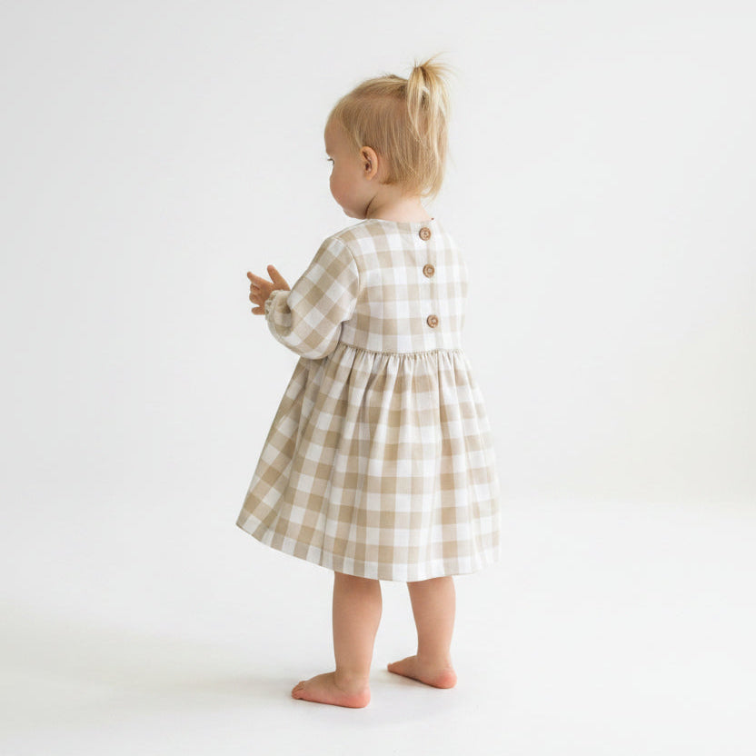A toddler's long-sleeve gingham dress in beige and white, featuring a buttoned neckline with three wooden buttons and a gathered skirt, laid flat on a white background next to a small sprig of baby's breath.