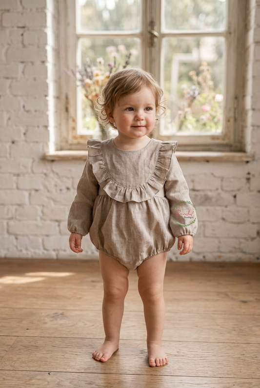A toddler girl wearing a light brown linen romper with ruffle details and a floral print on one sleeve, standing barefoot in a bright room with a brick wall and wooden floor.