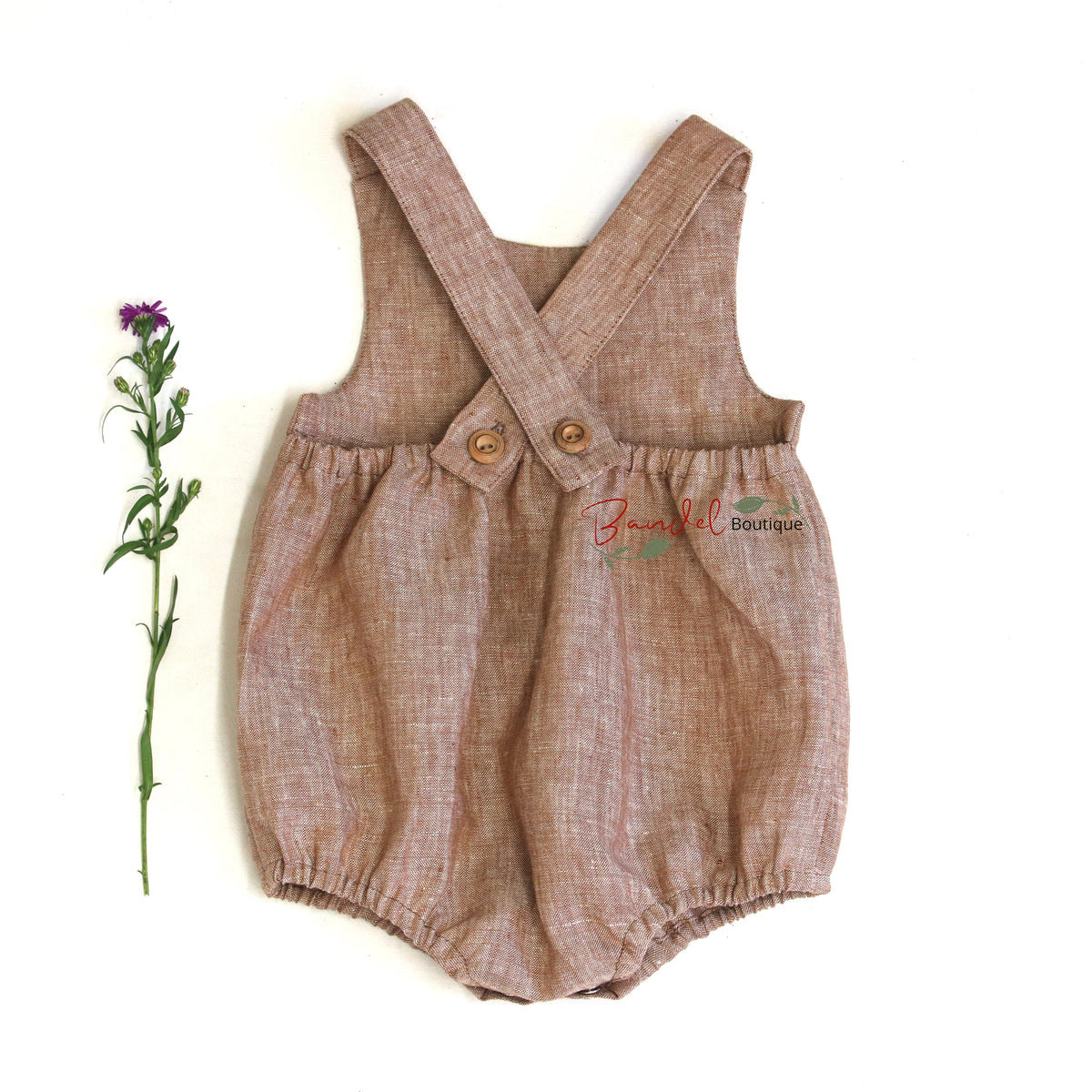 A light brown linen romper with adjustable shoulder straps and a button closure, laid flat on a white background next to a small purple flower stem. A brand watermark 'Fancy All Boutique' is visible on the right side.