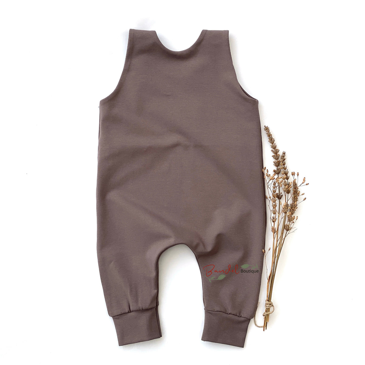 A sleeveless, one-piece baby romper in a muted taupe-brown color, laid flat on a white background next to a small bundle of dried flowers.
