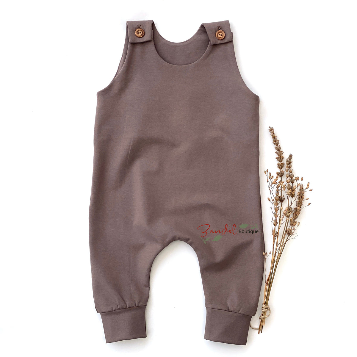 A sleeveless baby romper in a muted taupe-brown color, featuring adjustable shoulder straps with wooden buttons and elasticized leg cuffs. It is displayed flat on a white background next to a small dried floral sprig, with a 'Banzai Boutique' watermark near the bottom center.