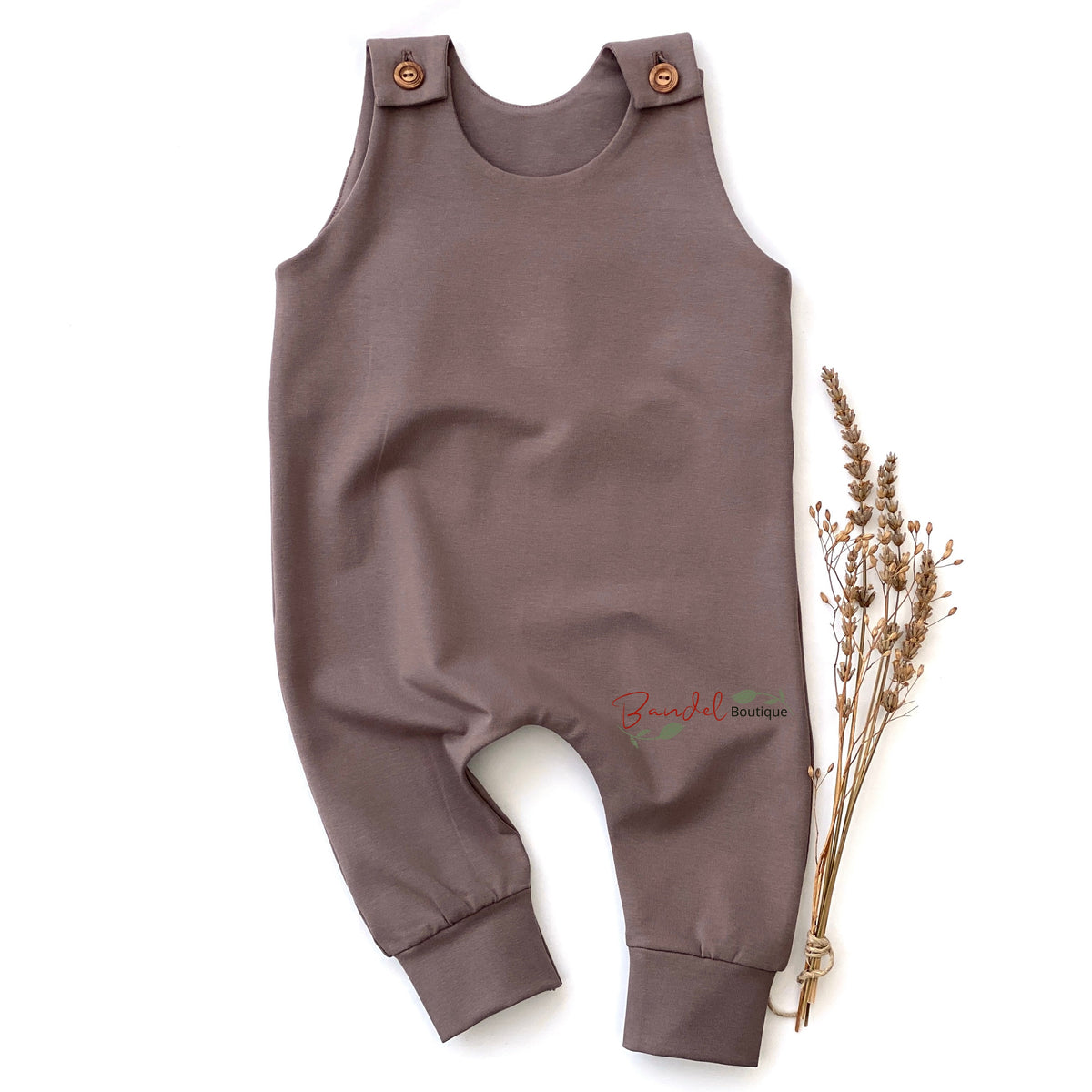 A sleeveless baby romper in a muted taupe-brown color, laid flat on a white background next to a small bundle of dried grass stems. The romper features adjustable shoulder straps with two brown buttons and cuffed ankles.