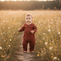 Child in a terracotta outfit standing on a path through a field of tall grass and wildflowers.