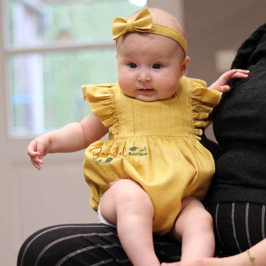 A baby wearing a mustard yellow ruffle-sleeve romper and matching bow headband sits on an adult's lap. The adult is wearing a black top and black-and-white pinstriped pants in a softly lit indoor setting.