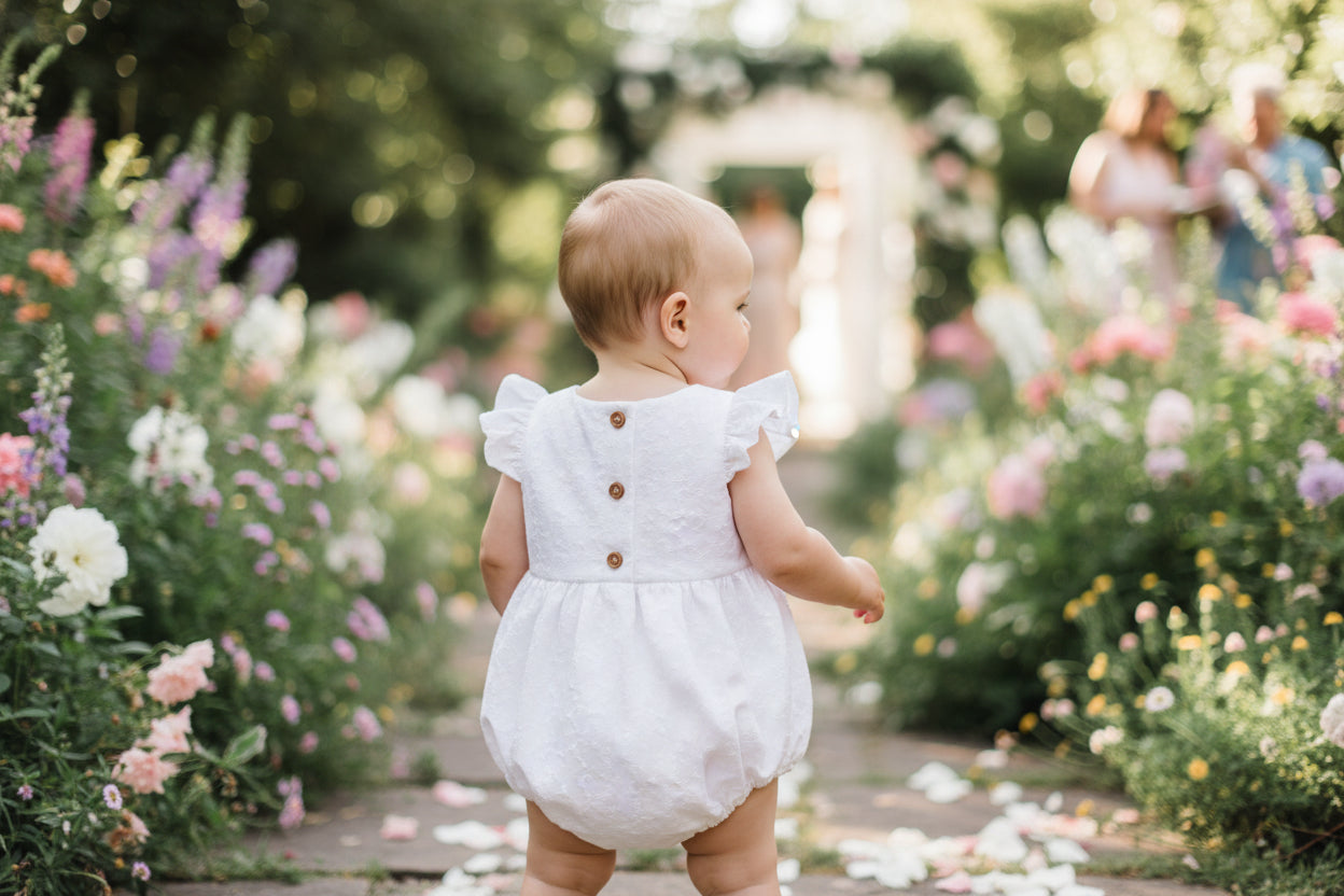 Baby in a white dress walking through a garden with flowers