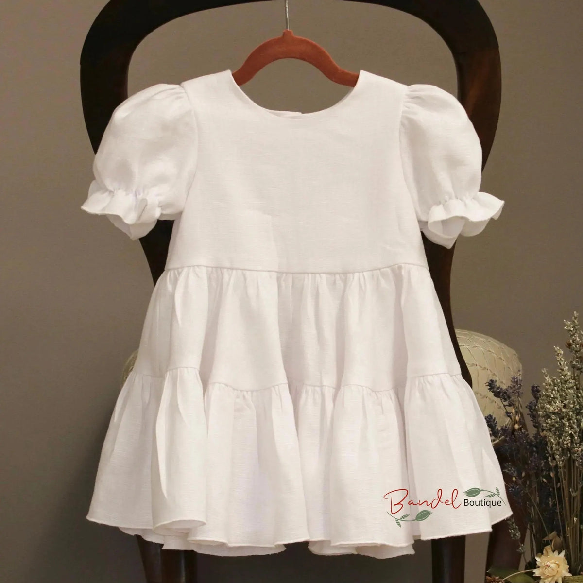 A white short-sleeve baby dress with puffed ruffled sleeves and a tiered skirt, hanging on a wooden hanger on a dark wooden chair.