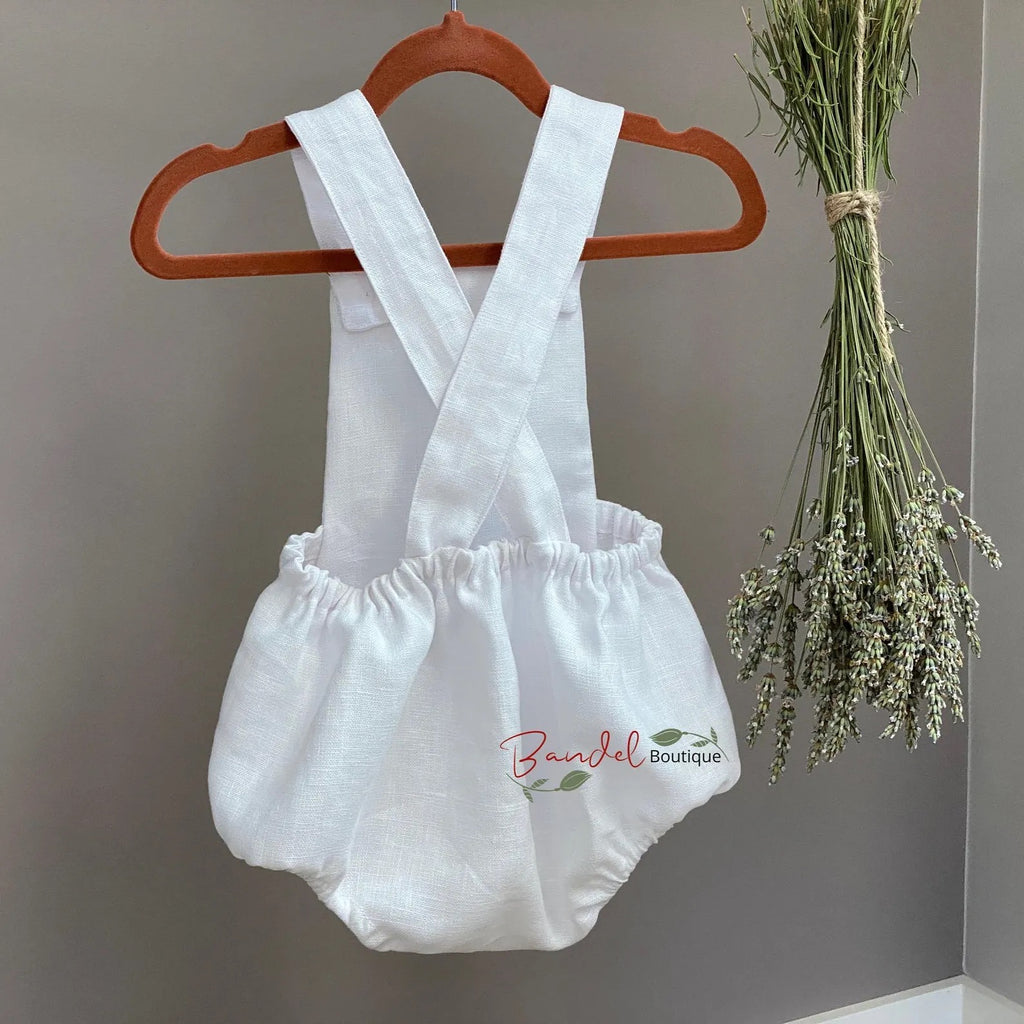 White baby romper with suspenders on a wooden hanger against a gray wall with dried flowers.