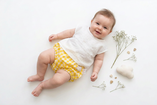A baby lying on a white background wearing a white sleeveless top and yellow-and-white gingham diaper cover, with small dried flowers and a soft white cloud-shaped prop nearby.