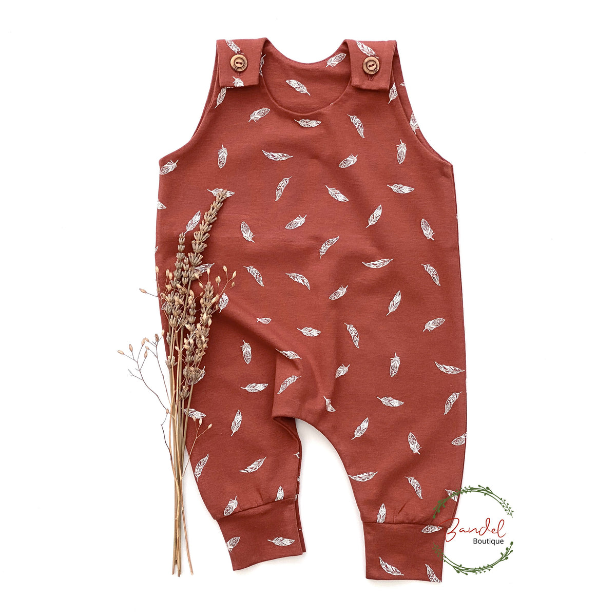 A rust-colored sleeveless baby romper with an all-over white feather print, styled flat-lay with dried grass stems on a white background.